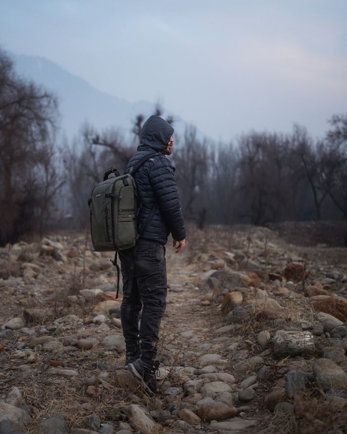 our-story A lone traveler with a backpack traversing a rocky path through a sparse forest, highlighting adventure and solitude.