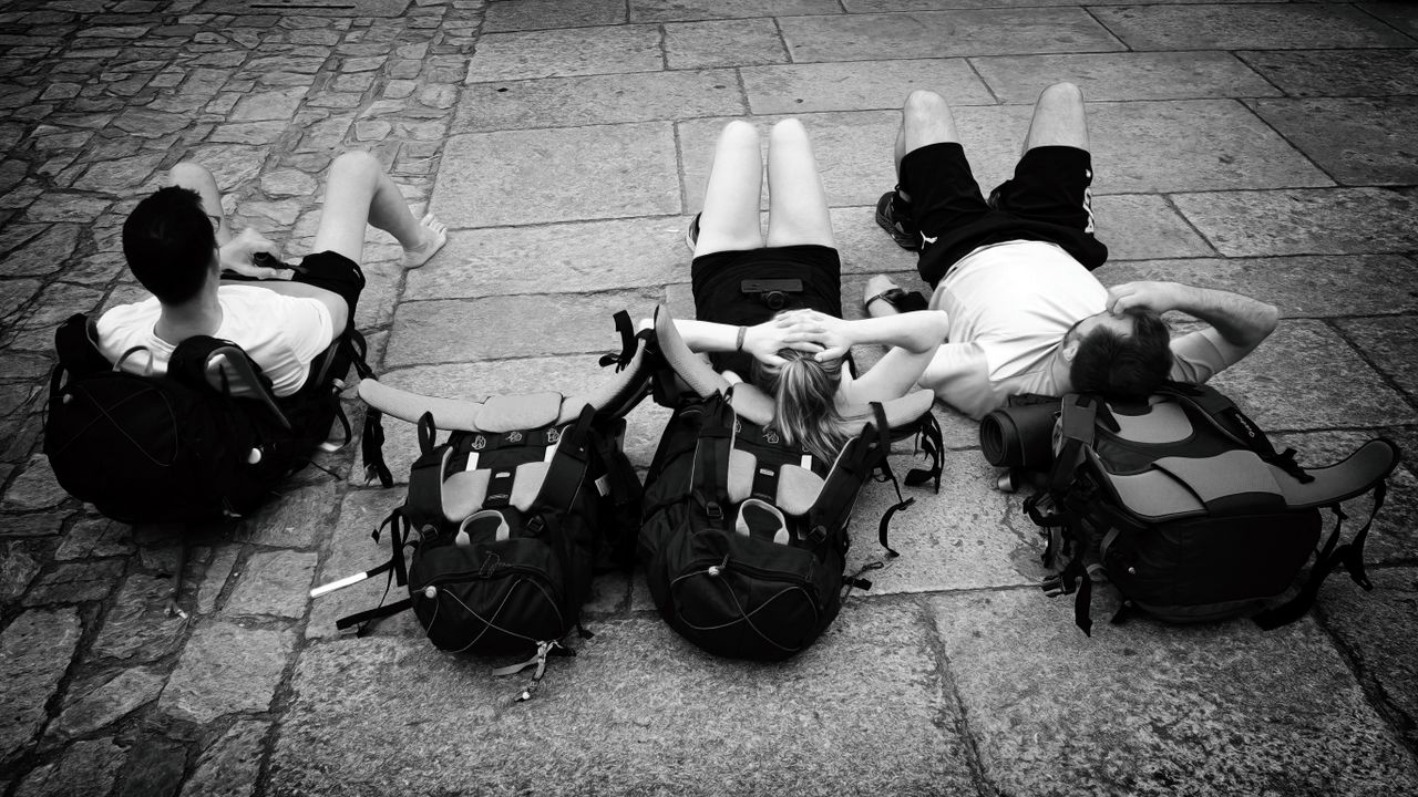 gallery-04 Three travelers resting with backpacks on a cobblestone street, embracing a moment of relaxation outdoors.
