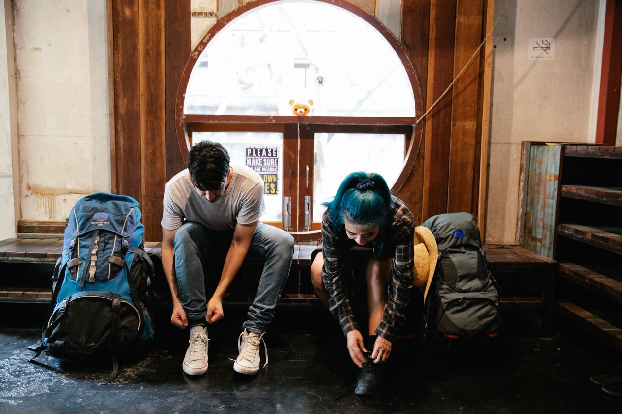 gallery-05 A man and woman sit indoors tying shoes next to backpacks, ready for travel.
