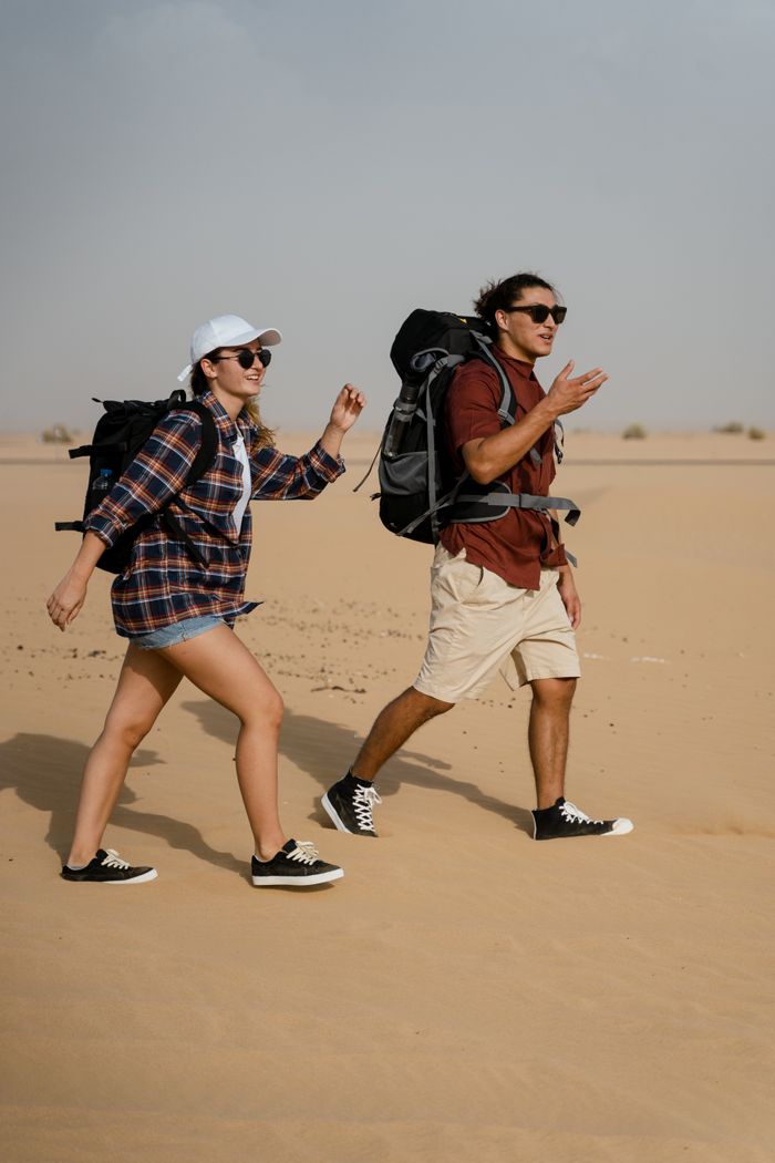gallery-03 Two young adults hiking with backpacks across an open desert on a sunny day, embracing adventure.
